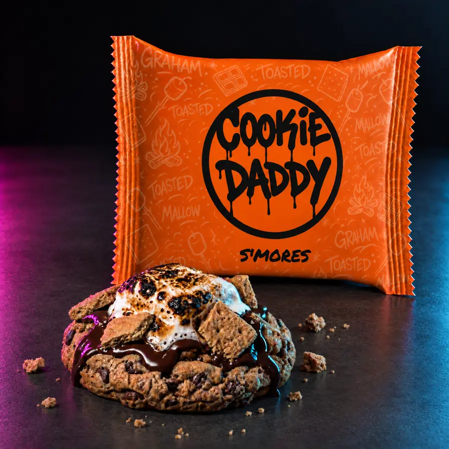 Neon orange Cookie Daddy wrapper, black ring tag with S'MORES printed under it, unwrapped s'mores cookie in front on dark concrete