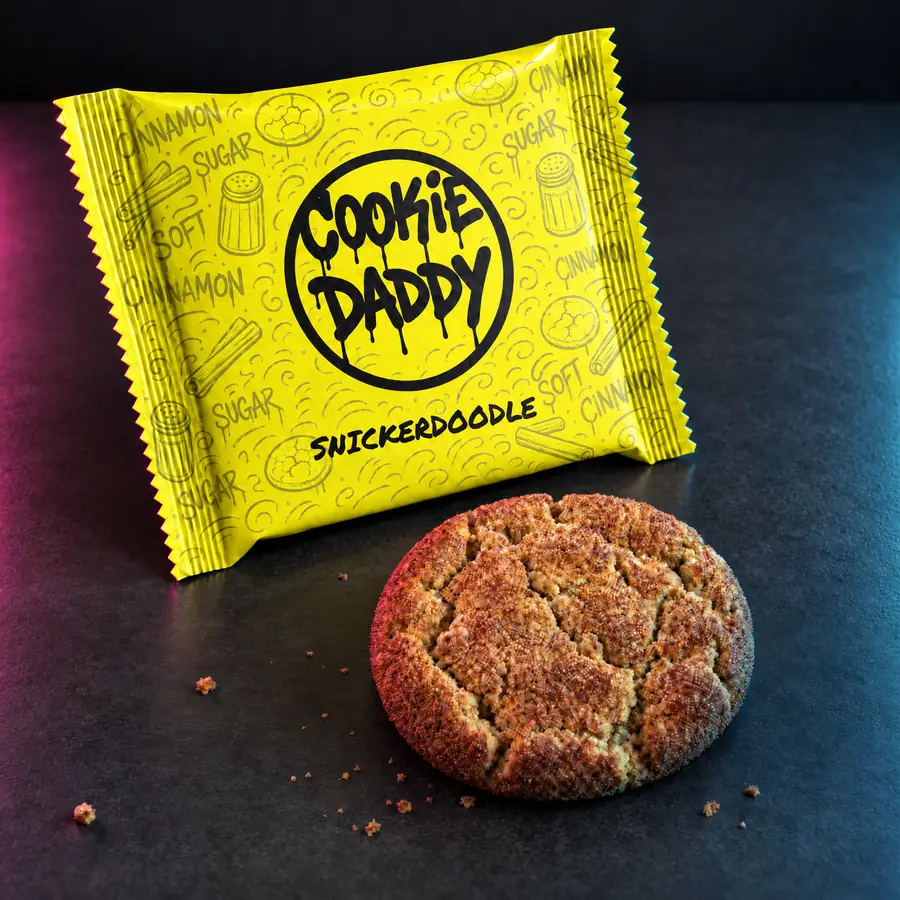 Neon yellow Cookie Daddy wrapper, black ring tag with SNICKERDOODLE printed under it, unwrapped snickerdoodle cookie in front on dark concrete